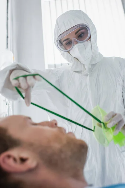 Doctor in hazmat suit placing oxygen mask on blurred patient in hospital — Stock Photo