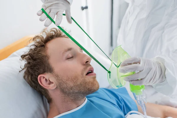 Médico en traje de materiales peligrosos y guantes de látex colocando máscara de oxígeno en el paciente enfermo en el hospital — Stock Photo