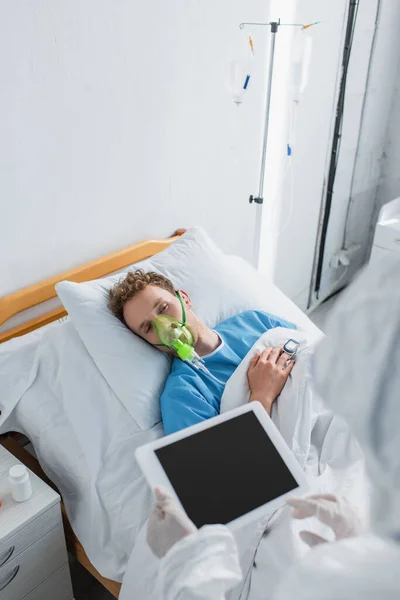 Високий кут зору хворого пацієнта в кисневій масці, який дивиться на лікаря в костюмі з туманом і латексними рукавичками з цифровим планшетом — стокове фото