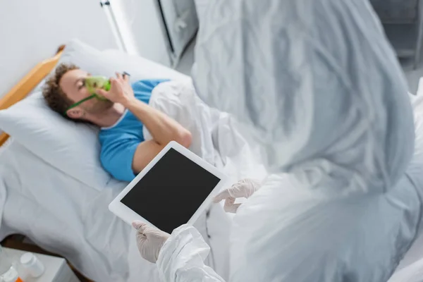 Vista de ángulo alto del médico en traje de materiales peligrosos y guantes de látex que sostienen la tableta digital con pantalla en blanco cerca del paciente enfermo - foto de stock