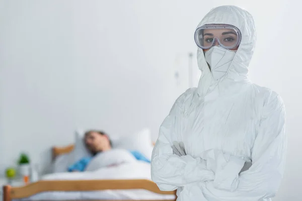 Medico in dispositivi di protezione individuale e occhiali in piedi con braccia incrociate vicino al paziente offuscato nel letto d'ospedale — Foto stock