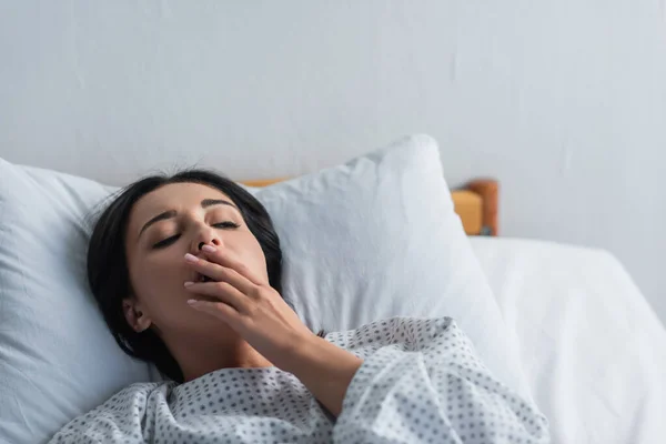 Брюнетка жінка в сукні пацієнта позіхання, лежачи в лікарняному ліжку — стокове фото