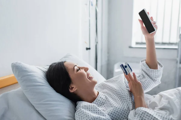 Happy woman with oximeter on finger using smartphone while lying on hospital bed — Stock Photo