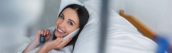 Улыбающаяся женщина с оксиметром на пальце разговаривает на смартфоне, лежа на больничной койке, баннер — стоковое фото