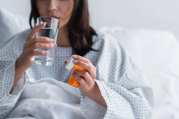 Cropped view of sick woman holding bottle with medication while taking pill in hospital — Stock Photo