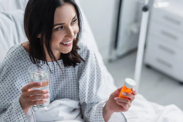 Smiling sick woman holding bottle with medication and glass of water in hospital — Stock Photo