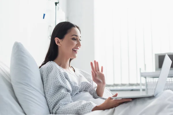 Alegre paciente saludando la mano durante tener videollamada en el hospital - foto de stock