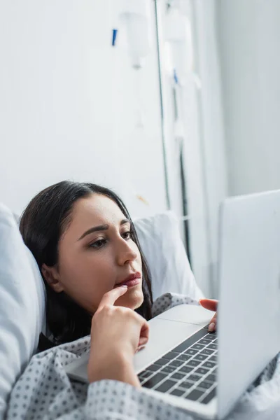 Фрілансер дивиться на ноутбук під час роботи віддалено в лікарняному ліжку — стокове фото