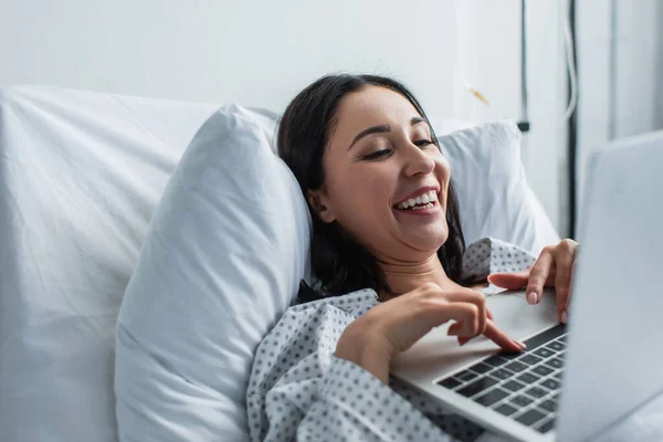 Mujer alegre viendo la película de comedia en el portátil en la cama del hospital - foto de stock