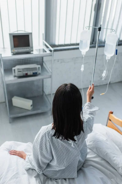 Visão traseira do paciente sentado na cama do hospital e de mãos dadas com frascos de terapia intravenosa — Fotografia de Stock