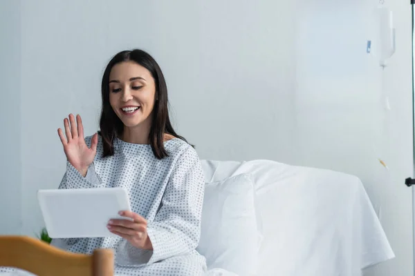 Mujer feliz en bata de paciente saludando la mano mientras tiene videollamada en el hospital - foto de stock