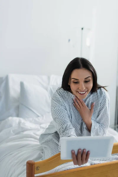 Mujer halagada en vestido de paciente sonriendo durante la videollamada en el hospital - foto de stock