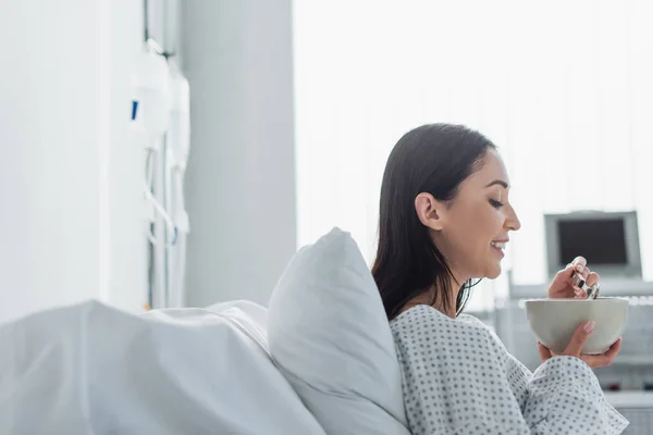 Vista laterale della donna allegra che tiene ciotola con colazione in ospedale — Foto stock