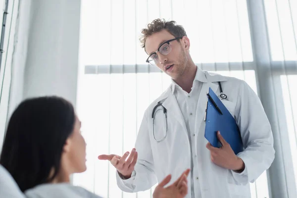 Médico rizado en gafas sujetando el portapapeles y haciendo gestos cerca del paciente - foto de stock