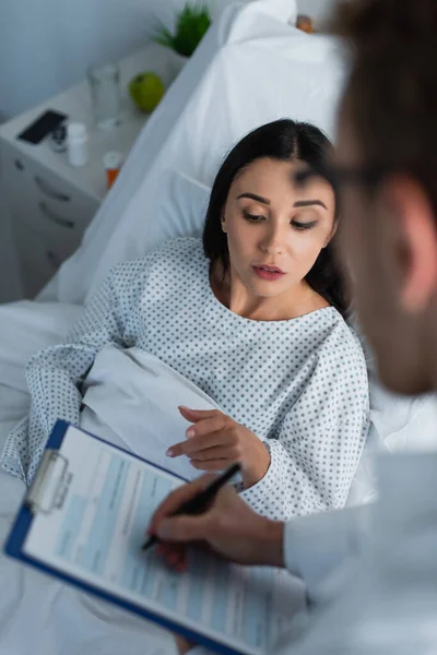 Hochwinkelaufnahme einer brünetten Frau, die auf Klemmbrett in der Nähe von verschwommenem Arzt zeigt — Stockfoto