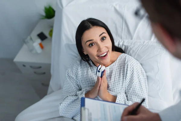 Vista de ángulo alto de la mujer sonriente con las manos orantes hablando con el médico borroso - foto de stock