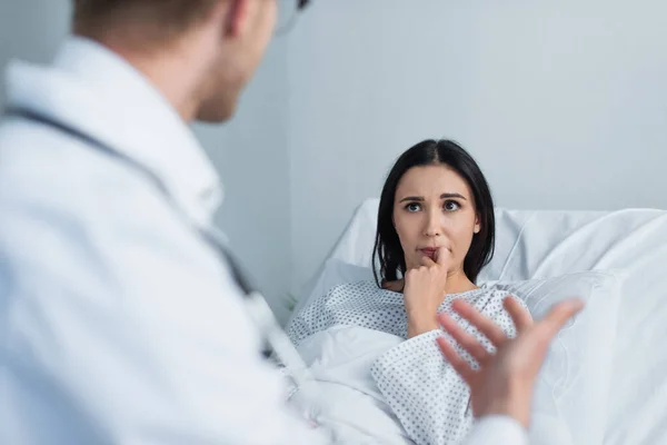 Брюнетка жінка в сукні пацієнта дивиться на розмитого лікаря в лікарні — стокове фото