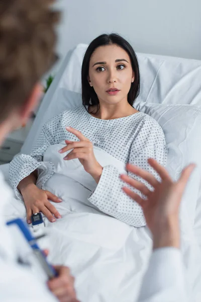 Morena mujer en vestido de paciente mirando borrosa médico en la clínica - foto de stock