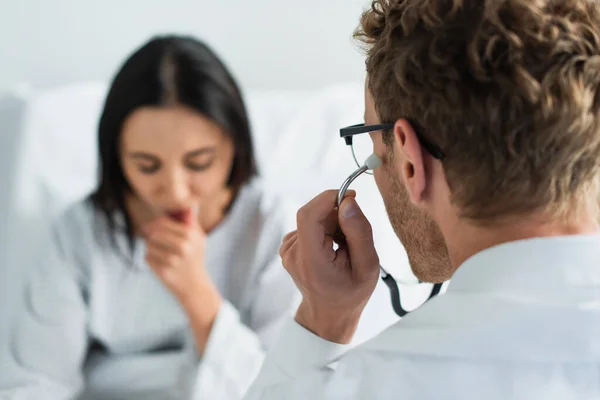 Doctor in glasses and stethoscope near blurred patient in hospital — Stock Photo