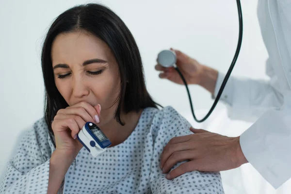 Sick woman in oximeter on finger coughing near blurred doctor with stethoscope — Stock Photo