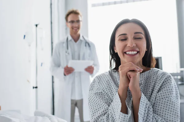 Femme gaie en robe de patient près du médecin flou avec tablette numérique mains serrées — Photo de stock