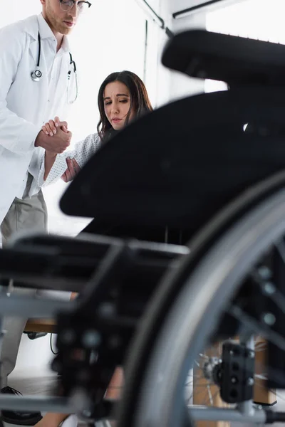 Médico en gafas de apoyo mujer cerca de la silla de ruedas borrosa - foto de stock