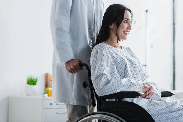 Médico de bata blanca de pie detrás de una mujer discapacitada sonriente en silla de ruedas - foto de stock