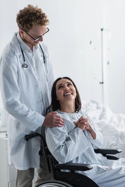 Médecin bouclé en manteau blanc debout derrière une femme handicapée souriante en fauteuil roulant — Photo de stock