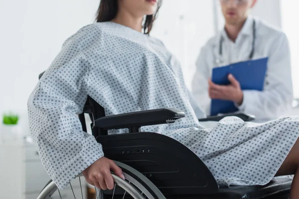 Vista recortada de la mujer discapacitada en silla de ruedas cerca del médico borroso - foto de stock