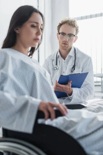 Doctor rizado sujetando portapapeles cerca de mujer discapacitada en silla de ruedas - foto de stock