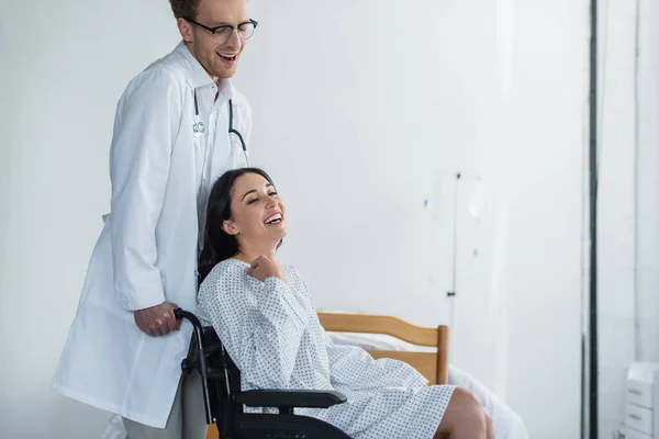Médico encaracolado em casaco branco em pé atrás de mulher deficiente alegre em cadeira de rodas — Fotografia de Stock