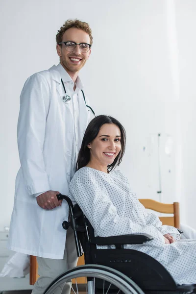 Allegro medico in camice bianco in piedi dietro gioiosa donna disabile in sedia a rotelle — Foto stock