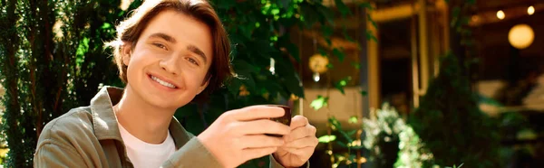 A young man in an olive green shirt smiles while savoring coffee in a trendy cafe setting, banner — Fotografia de Stock