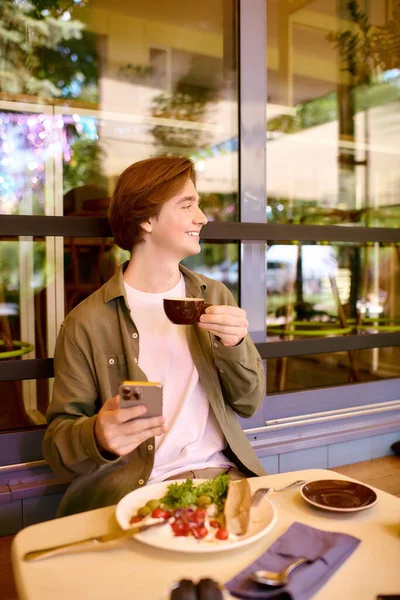 In a modern cafe, a young man sips coffee and smiles while enjoying his meal, embracing leisure. — Fotografia de Stock