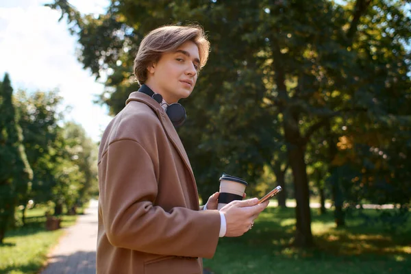 A young man in a fashionable coat strolls through an autumn park, sipping coffee. — Foto stock