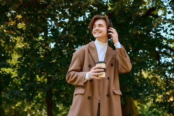 A young man in a fashionable coat smiles while relaxing outdoors in a vibrant autumn park. - foto de stock