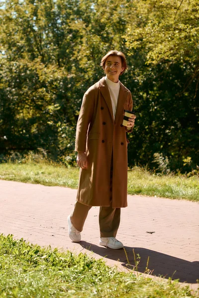 A young man walks cheerfully through an autumn park wearing a trendy coat, holding a drink. - foto de stock