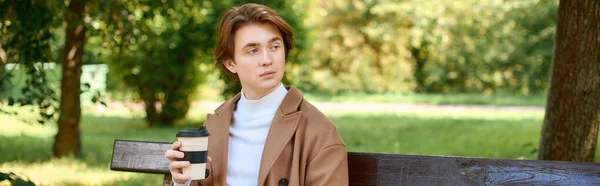 A young man sips a warm drink in a trendy coat at a park on a lovely autumn day, banner - foto de stock
