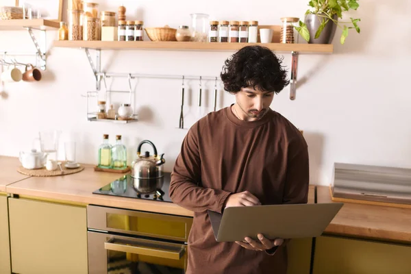 Handsome young man focuses on his laptop as he prepares a meal in a modern kitchen space. — Fotografia de Stock