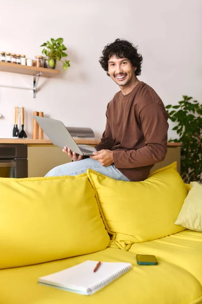 Happy young man sits on a vibrant yellow couch, using his laptop in a cozy living space. — стокове фото
