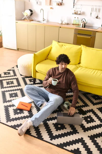 Handsome young man sits on the floor, sipping coffee and focused on his laptop in a bright room. — Stockfoto