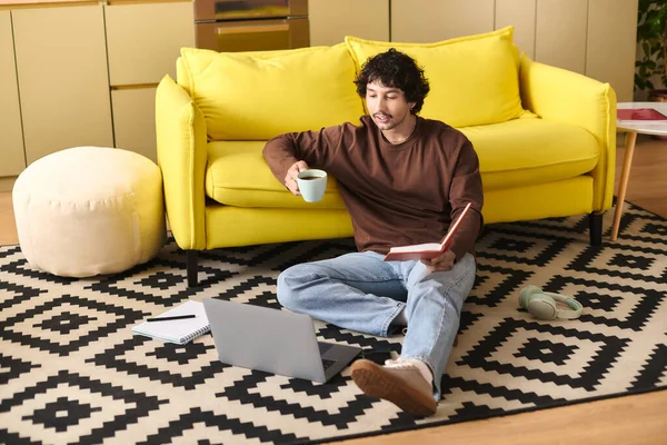 A handsome young man relaxes on the floor with a book and coffee in a sunny living space. - foto de stock
