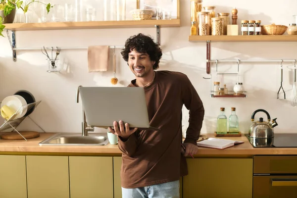 A young man stands in his modern kitchen, smiling as he uses his laptop. — стоковое фото