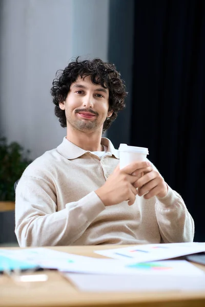 A young man with curly hair smiles while holding a coffee cup at his workspace. — стоковое фото