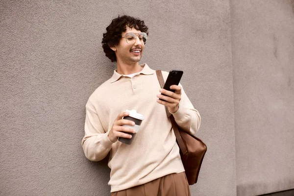 Handsome young man leans against a wall, smiling as he sips coffee and checks his phone. — Foto stock