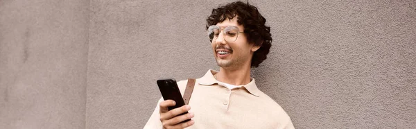 A stylish young man smiles as he interacts with his smartphone against a textured wall in the city. — стокове фото