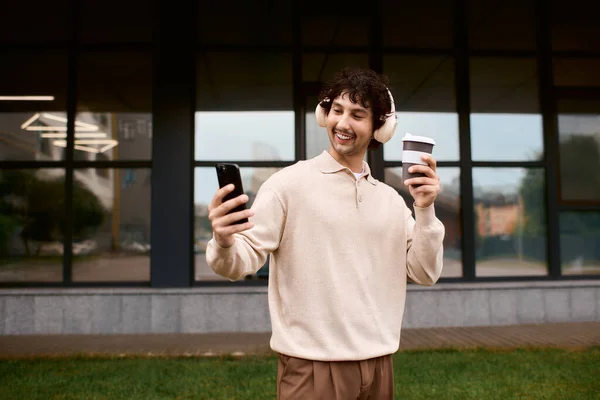 Handsome young man smiles as he takes a selfie, holding coffee and wearing headphones. — Foto stock
