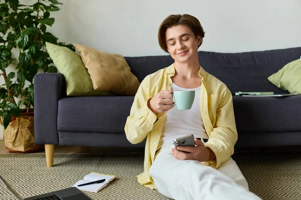 Jeune homme beau dans une chemise jaune assis sur le sol, sirotant du café et utilisant son téléphone. — Photo de stock