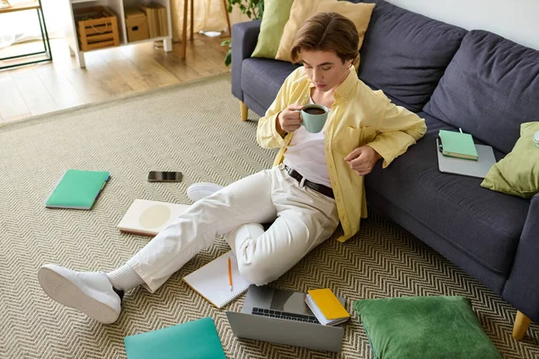 Fashionable young man sits on the floor, sipping coffee and focused on his laptop in a stylish room. — Stock Photo