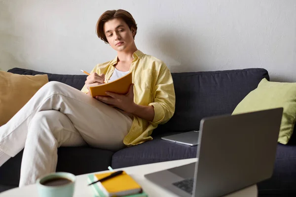 Young fashionable man sits comfortably on a sofa, jotting down notes while engaged with his laptop. — Stock Photo